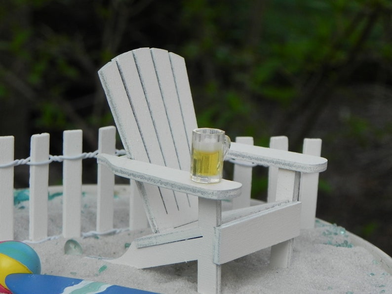 May include: A white wooden adirondack chair with a small glass of beer on the armrest. The chair is on a white sandy beach with a white picket fence in the background.