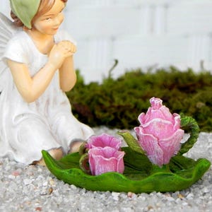 May include: A fairy figurine in a white dress and green head covering kneels near a miniature tea set. The tea set includes a pink teapot and two cups, all shaped like roses, on a green leaf-shaped tray.