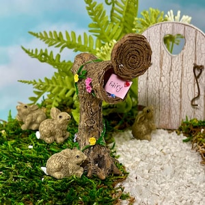 May include: A miniature fairy garden scene featuring a brown tree trunk mailbox with a pink letter that says "fairy". Several small brown bunny figurines are scattered around the mailbox. A white door with a heart-shaped cutout is in the background.