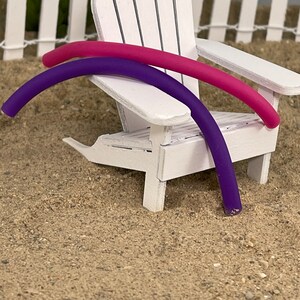 May include: Two pool floats, one purple and one pink, are leaning against a white wooden chair. The chair is sitting on a sandy surface.