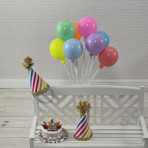 May include: A miniature birthday scene with a small cake, party hats, and colorful balloons. The cake is on a silver platter and topped with flowers. The party hats have rainbow stripes and gold trim. The balloons are in various colors.