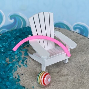 May include: A white wooden Adirondack chair with a pink pool noodle resting on the armrest. A colourful beach ball sits on the sand below the chair.