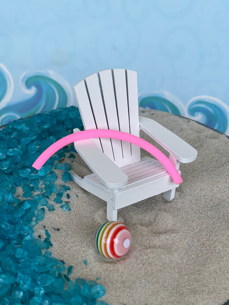 May include: A white wooden Adirondack chair with a pink pool noodle resting on the armrest. A colourful beach ball sits on the sand below the chair.