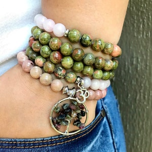 May include: A stack of bracelets made with green, pink, and white stones. The bracelets are layered on a person's wrist and feature a tree of life charm with a small Om symbol.