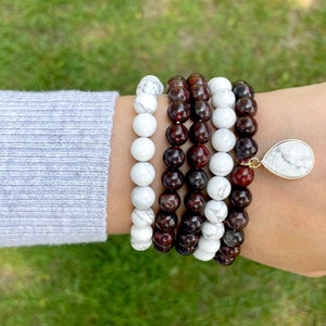May include: A set of four bracelets, two are white and two are dark red. The bracelets are made of round beads and are stacked on a person's wrist. A white teardrop-shaped pendant hangs from the bottom of the bracelets.