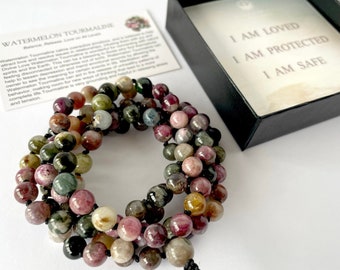 May include: A multi-coloured watermelon tourmaline gemstone mala necklace with black string. The necklace is in a small black box with the words "I am loved, I am protected, I am safe" printed inside.