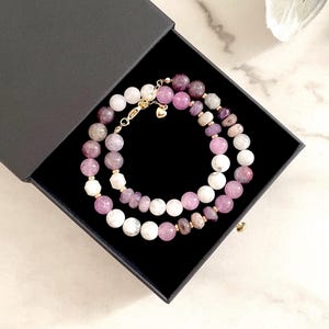 May include: Two beaded necklaces in a black box. The necklaces feature white, pink, and purple beads. The box is open, revealing the necklaces inside. The necklaces are made of round beads and are connected by gold-colored clasps.