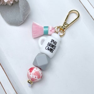 Może przedstawiać: Brelok do kluczy ze złotym zapięciem, różową chwostą i wzorem z koralików. Koraliki obejmują biały kubek z napisem "SMALL BUSINESS OWNER", szary geometryczny koralik oraz różowo-biały koralik we wzory. Brelok leży na białej powierzchni.