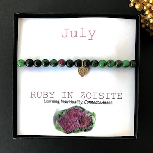 May include: A black box with a white card inside. The card has the word "July" printed in pink and the words "RUBY IN ZOISITE" printed in black. Below the words is the text "Learning, Individuality, Connectedness". There is a small green and pink stone in the bottom right corner of the card. A green, black, and pink beaded bracelet with a gold heart charm is on top of the card.