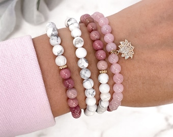 May include: A set of four bracelets featuring white howlite, pink rose quartz, and pink rhodochrosite beads. The bracelets are stacked on a wrist and include a gold lotus charm with clear rhinestones.