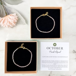 May include: Two small boxes with pink opal bracelets inside. The boxes are open and the bracelets are visible. A card with the text "OCTOBER Pink Opal I LOVE AND ACCEPT MYSELF UNCONDITIONALLY" is in front of the boxes.