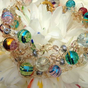 May include: A gold wire bracelet with colorful glass beads, including blue, green, pink, and clear beads. The bracelet is on a white flower with colorful paint splatters.