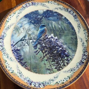 Könnte beinhalten: Ein runder dekorativer Porzellanteller mit einer zentralen Abbildung eines Blauvogels, der auf einem Ast mit violetten Glyzinienblüten sitzt. Der Rand ist cremefarben mit einer Bordüre aus kleinen weißen Blüten und einem goldfarbenen Rand. Die Marke Lenox ist sichtbar.