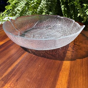 May include: Clear glass leaf-shaped bowl with textured detailing. The bowl's design mimics the veins of a leaf. The bowl is sitting on a wooden surface.