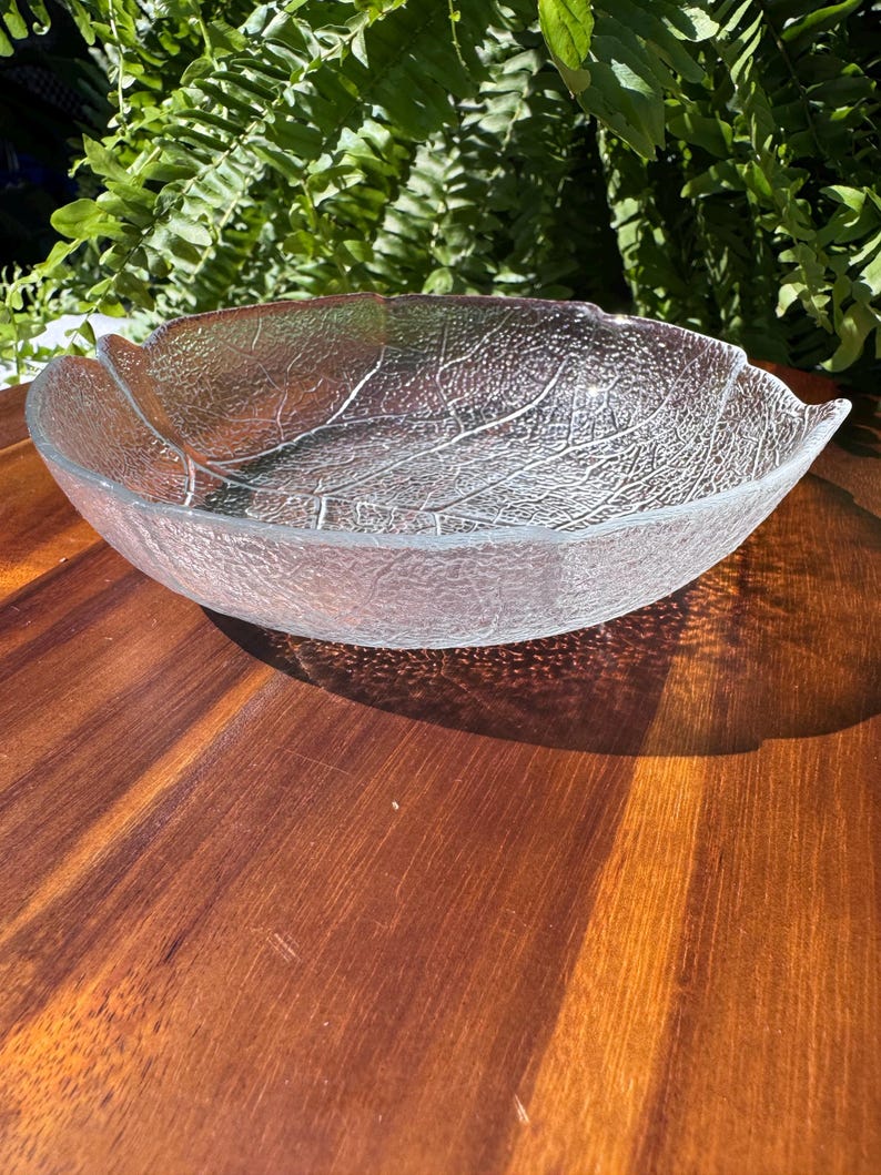May include: Clear glass leaf-shaped bowl with textured detailing. The bowl's design mimics the veins of a leaf. The bowl is sitting on a wooden surface.