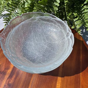 May include: Clear glass serving bowl with a leaf design. The bowl has a textured surface and a scalloped edge, and is sitting on a wooden surface. Ferns are in the background.