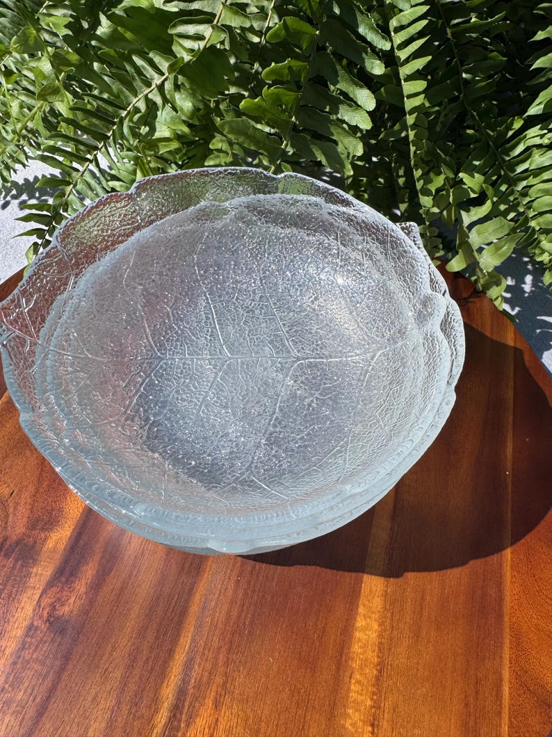 May include: Clear glass serving bowl with a leaf design. The bowl has a textured surface and a scalloped edge, and is sitting on a wooden surface. Ferns are in the background.