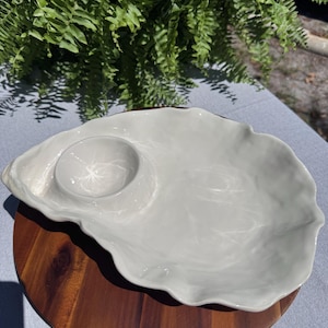 May include: A light gray ceramic oyster-shaped serving platter with a small circular dip bowl. The platter sits on a round wooden board. Ferns are visible in the background.