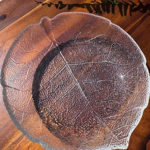 May include: A clear glass leaf-shaped bowl with detailed vein patterns. The bowl sits on a wooden surface, with a natural leaf design. The bowl's edges are slightly raised, and the overall design is organic and decorative.