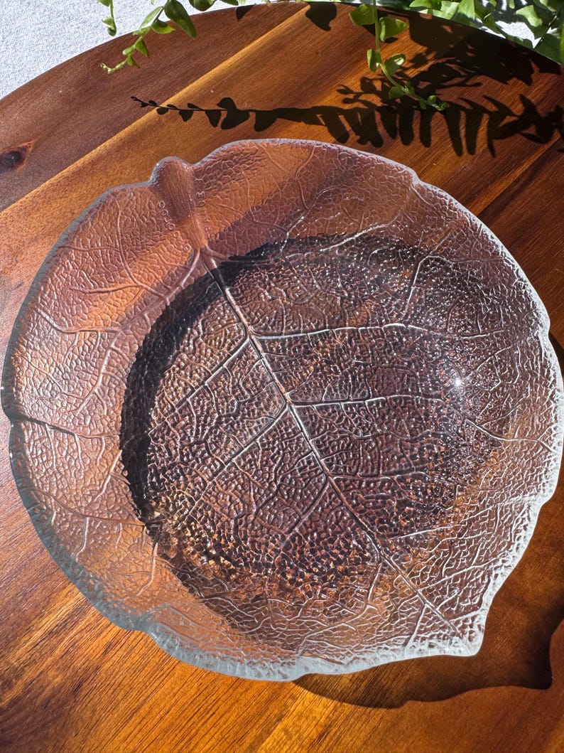 May include: A clear glass leaf-shaped bowl with detailed vein patterns. The bowl sits on a wooden surface, with a natural leaf design. The bowl's edges are slightly raised, and the overall design is organic and decorative.