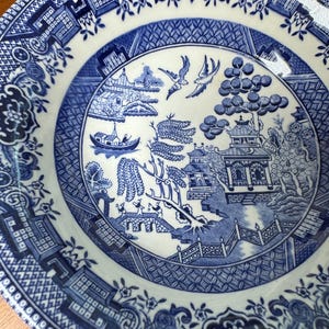 May include: A blue and white ceramic plate with a detailed Willow pattern design. The plate features a central scene with a pagoda, birds, and a boat, surrounded by a decorative border. The plate is round and has a glossy finish.