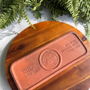 Puede incluir: Un ladrillo rectangular de terracota para panera con esquinas redondeadas. La superficie presenta el texto en relieve BREAD BASKET BRICK, USE WITH EXTREME CARE WHEN HOT y un logotipo circular central de Longaberger Pottery.