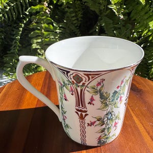 May include: A white ceramic mug featuring a decorative pattern of trailing green vines with small pink and red flowers. The design includes vertical brown and gold geometric columns between the floral motifs, with a gold rim around the top edge.