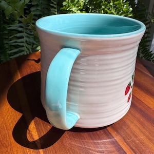 May include: A ceramic mug with a light blue handle and interior. The exterior is white with a textured, ribbed design and a small strawberry illustration. The mug is sitting on a wooden surface.