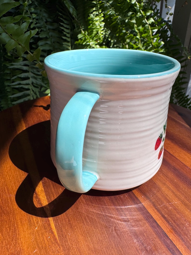 May include: A ceramic mug with a light blue handle and interior. The exterior is white with a textured, ribbed design and a small strawberry illustration. The mug is sitting on a wooden surface.