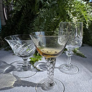 May include: Three clear crystal stemmed glasses with intricate designs. One glass has a wide bowl and a twisted stem, another has a diamond pattern, and the third is a tall, slender glass. The glasses are arranged on a light gray surface.