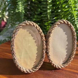 May include: Two oval-shaped picture frames featuring a textured, rope-like border in a metallic gold finish. Each frame contains a plain, off-white insert, and the frames are positioned side-by-side, displaying a consistent design and structure.