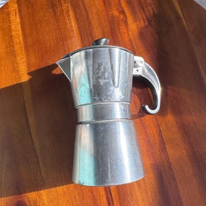 May include: A silver-colored aluminum moka pot coffee maker featuring an octagonal upper chamber, a tapered base, and a black plastic handle with a matching black knob on the lid. The metal surface shows signs of wear and light surface scratches.