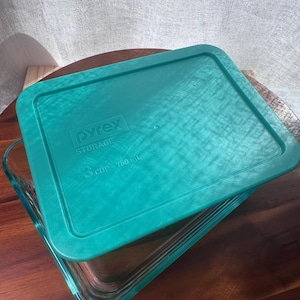 Puede incluir: Una fuente de horno Pyrex de vidrio transparente con una tapa de color turquesa. La tapa rectangular tiene el logotipo de Pyrex Storage y la capacidad de 750 ml. La fuente está sobre una superficie de madera.