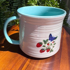 May include: White ceramic mug with a light blue interior and handle. The mug features a design of red strawberries and a blue butterfly. The mug has a ridged texture and is sitting on a wooden surface.