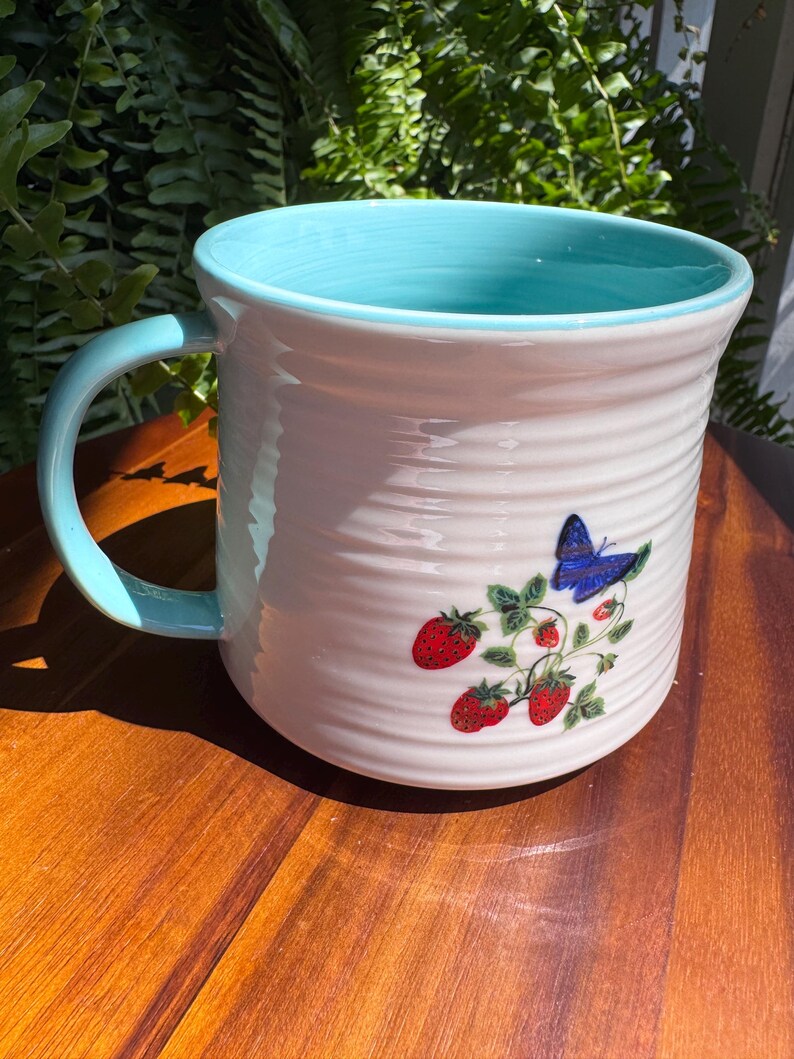 May include: White ceramic mug with a light blue interior and handle. The mug features a design of red strawberries and a blue butterfly. The mug has a ridged texture and is sitting on a wooden surface.