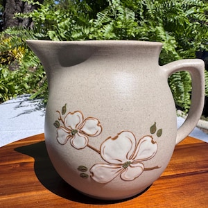 May include: A beige ceramic jug with a handle and a spout. The jug is decorated with white and brown floral designs. The jug is sitting on a wooden surface.