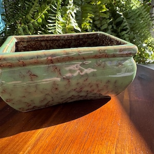 May include: A rectangular ceramic planter featuring a light green glaze with a mottled, dark brown crackle pattern throughout the exterior and interior surfaces. The vessel has a wide, open top with a slightly flared rim and rounded corners.