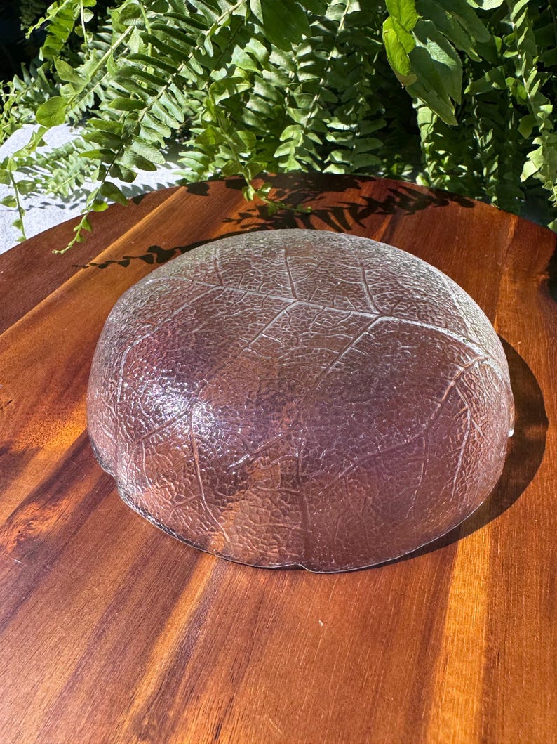 May include: A clear glass bowl with a leaf pattern. The bowl is round and has a textured surface. The bowl is sitting on a wooden surface.