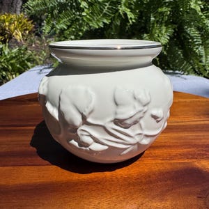 Peut inclure: Un vase en céramique blanc avec un bord argenté et un motif floral en relief. Le vase rond repose sur une surface en bois, avec une toile de fond de feuillage vert. Le vase est un objet décoratif.