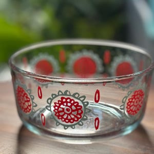 May include: Clear glass bowl with red and gray floral designs. The bowl has an oval shape and sits on a wooden surface. The floral pattern includes red flower-like shapes with gray outlines and teardrop accents.