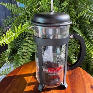 Könnte beinhalten: Eine French Press Kaffeebereiter aus klarem Glas mit schwarzem Kunststoffrahmen, Griff und Deckel. Ein Metallkolben mit schwarzem Knopf befindet sich oben. Ein rotes Etikett am Glaskörper zeigt den weißen Text original FRENCH PRESS in einem Rechteck.