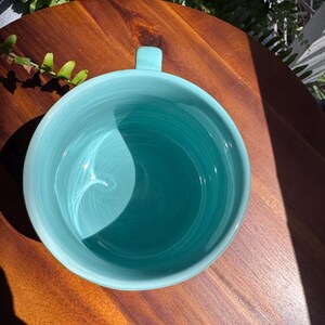 May include: A turquoise ceramic mug with a rounded shape and a small handle. The mug is set on a dark wooden surface, with natural light casting shadows. The mug's interior is a lighter shade of turquoise.