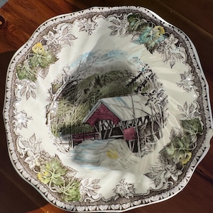 May include: A round ceramic plate with a scalloped edge featuring a central illustration of a red covered bridge in a snowy landscape. The border is decorated with a brown and green botanical pattern of leaves and flowers on a cream background.
