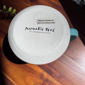 May include: White ceramic mug with a turquoise handle, bottom facing up. The mug has the text "Nathalie Lete for Anthropologie" and a sticker that reads "Dishwasher & Microwave Safe Made in China/Fabriqu&eacute; en Chine."