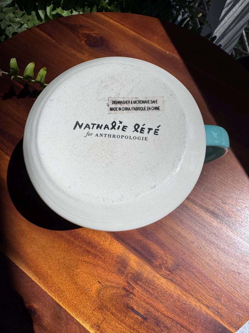 May include: White ceramic mug with a turquoise handle, bottom facing up. The mug has the text "Nathalie Lete for Anthropologie" and a sticker that reads "Dishwasher & Microwave Safe Made in China/Fabriqu&eacute; en Chine."
