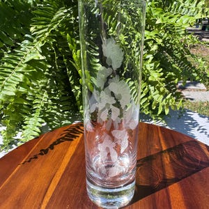 May include: A clear glass vase with etched floral designs. The cylindrical vase is tall and slender, with a slightly flared base. The etched design features leaves and flowers. The vase is sitting on a wooden surface, with green foliage in the background.
