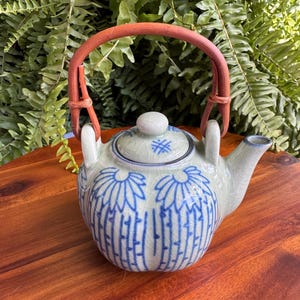 May include: A ceramic teapot with a light green and blue floral design. The teapot has a brown handle and a matching lid. The teapot is sitting on a wooden surface.