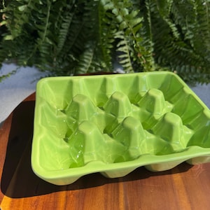 Puede incluir: Un portahuevos de cerámica verde brillante con acabado brillante. La huevera tiene forma rectangular con bordes redondeados y compartimentos individuales. Se encuentra sobre una superficie de madera.