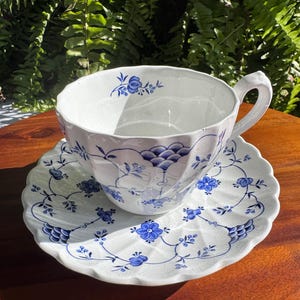 Puede incluir: Un juego de taza de té y platillo de cerámica blanca con borde festoneado y un delicado patrón floral azul. La superficie de ambas piezas presenta un acabado de esmalte craquelado, con enredaderas azules, pequeñas flores y motivos en abanico.