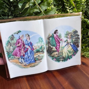 May include: A white ceramic decorative object shaped like an open book with gold-toned edges. The two pages feature circular illustrations of 18th-century figures in colourful period clothing, including pink coats and blue dresses, set in outdoor landscapes.